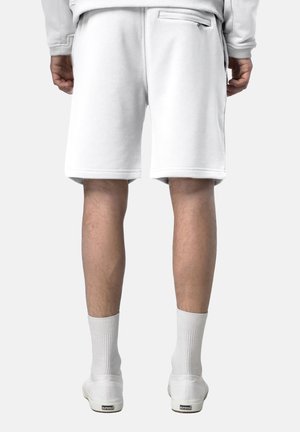 Uomo che indossa pantaloncini bianchi fino al ginocchio, calzini bianchi e scarpe slip-on bianche, in piedi con la schiena rivolta in avanti.