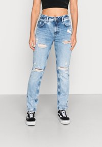 Jeans de cintura alta, rasgados e de um azul claro, com múltiplos remendos desgastados e um design de perna reta, combinados com ténis pretos e brancos.