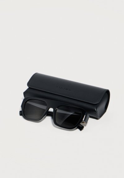 Μαύρα ορθογώνια γυαλιά ηλίου με σκούρους φακούς τοποθετημένα μπροστά από μια αντίστοιχη μαύρη δερμάτινη θήκη σε ανοιχτό φόντο.