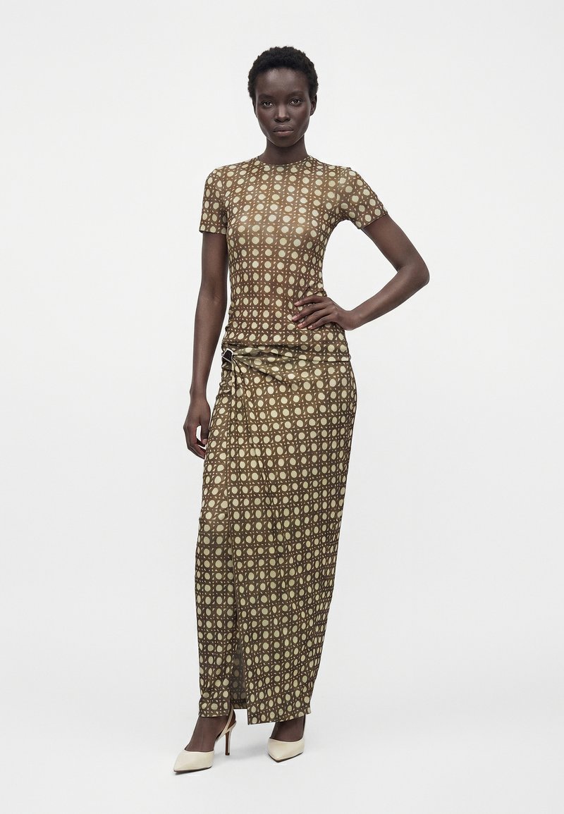 Femme aux cheveux courts portant un haut marron ajusté à motifs et une longue jupe assortie avec une fente à l'avant, debout, une main sur la hanche.