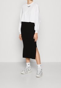 Weißes, kurzes Langarmshirt, schwarze, gerippte Midirock mit Seitenschlitz, kombiniert mit weißen Sneakers mit schwarzen Akzenten und Logo.