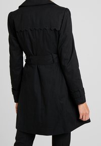 Czarny płaszcz trencz z ozdobnym karczkiem w kształcie fali, paskiem w talii i usztywnionymi rękawami; wykonany z gładkiej tkaniny o minimalistycznym wzornictwie.