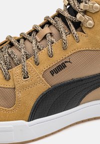 Närbild av en tan och svart Puma-sneaker med mönstrade snören, material av mocka och mesh, metallöglor och vit sula.