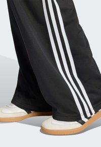Black athletic pants with white side stripes, made of smooth fabric. Paired with white sneakers featuring gum sole and black accents.