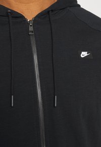 Svart zip-up hoodie i strukturerad tyg, med en liten Nike-logga på bröstet och dragsnören med plastspetsar.