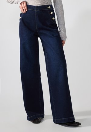 Woman wearing high-waist wide-leg dark denim pants with white buttons on pockets and seam details, paired with grey long-sleeve top and black shoes.