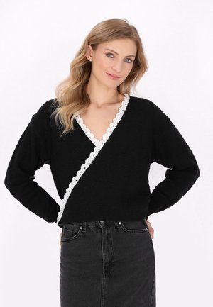 Schwarzer Wickelsweater mit einem gewellten weißen Saum entlang des Halsausschnitts, ausgestattet mit langen Ärmeln und einem strukturierten Strickmuster. Kombiniert mit einem schwarzen Rock.