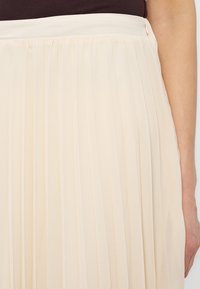 Jupe plissée couleur crème portée avec un haut marron foncé rentré à l'intérieur, montrant la taille et le bras droit de la personne.