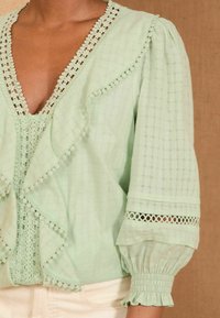 Kobieta w jasnozielonej bluzce z koronkowym wykończeniem, falbaniastym przodem oraz bufiastymi rękawami z elastycznymi mankietami na brązowym tle.