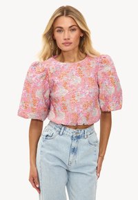 Crop top floral rose avec manches bouffantes, présentant une texture smockée et des accents orange vif. Associé à un jean en denim clair.