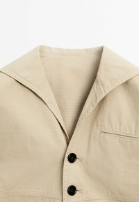 Beige collared jacket with wide lapels, two black buttons, and a small horizontal chest pocket on the right side.