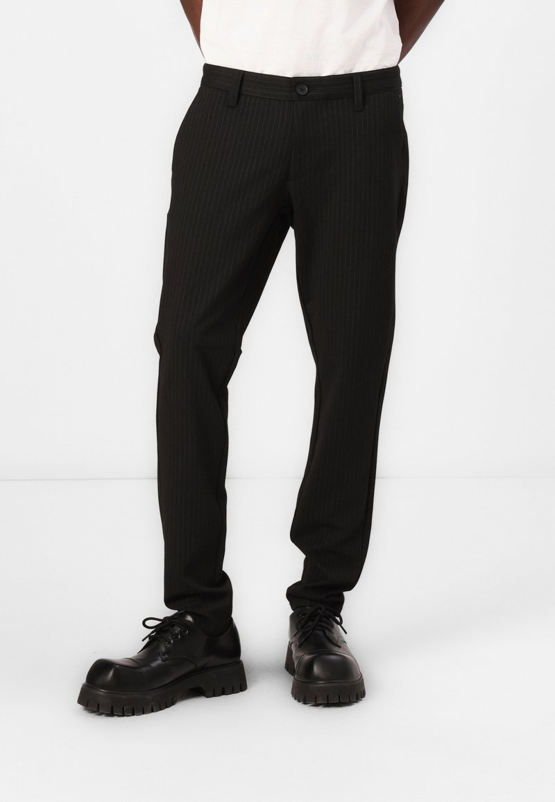 Pantalon noir à rayures avec une coupe droite, dotée d'une fermeture à boutons, de passants de ceinture et de poches, associé à des chaussures noires à semelles épaisses.