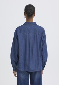 Chemise en denim bleu foncé, avec un col pointu, des manches longues à poignets boutonnés et un détail de couture au dos.