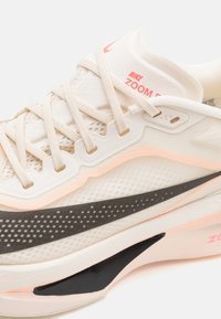 Nike Zoom Fly løbesko i lys beige, med en mesh-overdel, tonale snørebånd og en sort og pink accent på sålen. Struktureret overflade med mønstre.