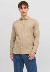 Light brown long-sleeve shirt made of cotton, featuring a collar, button front, and straight cut. Worn with blue denim jeans.