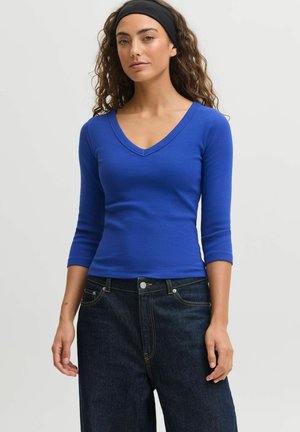 Kvinde med krøllet hår iført sort pandebånd, tætsiddende blå V-hals top med trekvartærmer og mørkeblå højttaljede jeans.