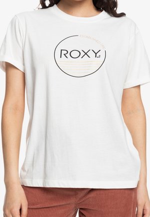 Vrouw draagt een wit T-shirt met korte mouwen met een rond "ROXY"-logo en lichtbruine lijnen, gecombineerd met roestkleurige broek.