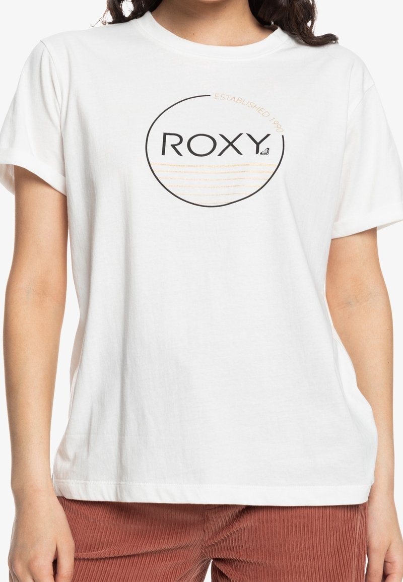 Femme portant un t-shirt blanc à manches courtes avec un logo circulaire "ROXY" et des lignes brun clair, associé à un pantalon couleur rouille.