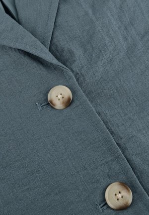 Tissu gris-bleu avec deux grands boutons beige attachés, montrant un tissage texturé et des couches de vêtements superposées.