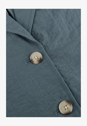 Modro-siva tkanina z dvema velikima bež gumboma, pritrjenima, ki prikazuje teksturirano tkanje in prekrivajoče se plasti oblačila.