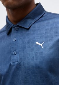 Κοντινό πλάνο ενός ατόμου που φοράει μπλε πόλο πουκάμισο με μοτίβο, με τρία κουμπιά και λευκό λογότυπο Puma στο στήθος.
