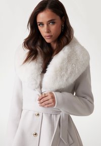 Manteau en mélange de laine couleur blanc cassé avec un col en fausse fourrure, des boutons dorés et une ceinture à la taille. Texture lisse avec une coupe ajustée.