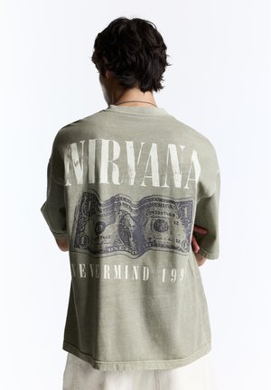 Personne aux cheveux foncés portant un t-shirt vert délavé du groupe Nirvana avec un motif billet de dollar et le texte "Nevermind 1991" au dos.