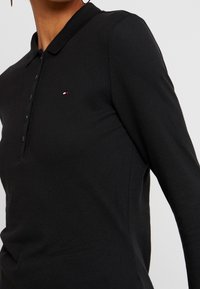 Mustapaitakäsi polo-paita, joka on valmistettu teksturoidusta kankaasta. Paita sisältää kauluksen, nappilistan ja pienen logoembleemin punaisessa, valkoisessa ja sinisessä värissä.