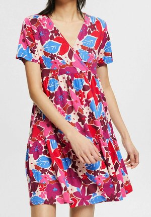 Frau trägt ein kurzärmeliges Kleid mit V-Ausschnitt und geraffter Taille, das ein lebendiges Blumenmuster in Rot-, Blau-, Rosa- und Lilatönen zeigt.