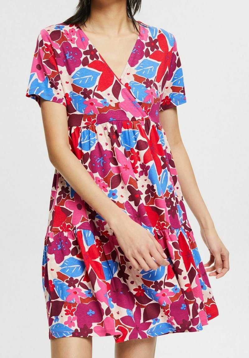 Femme portant une robe à manches courtes et col en V avec une taille froncée, présentant un motif floral vibrant aux tons rouges, bleus, roses et violets.
