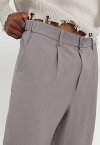 Gray pleated trousers made of smooth fabric with a button closure and slanted front pockets. The waistband is flat with subtle stitching details.