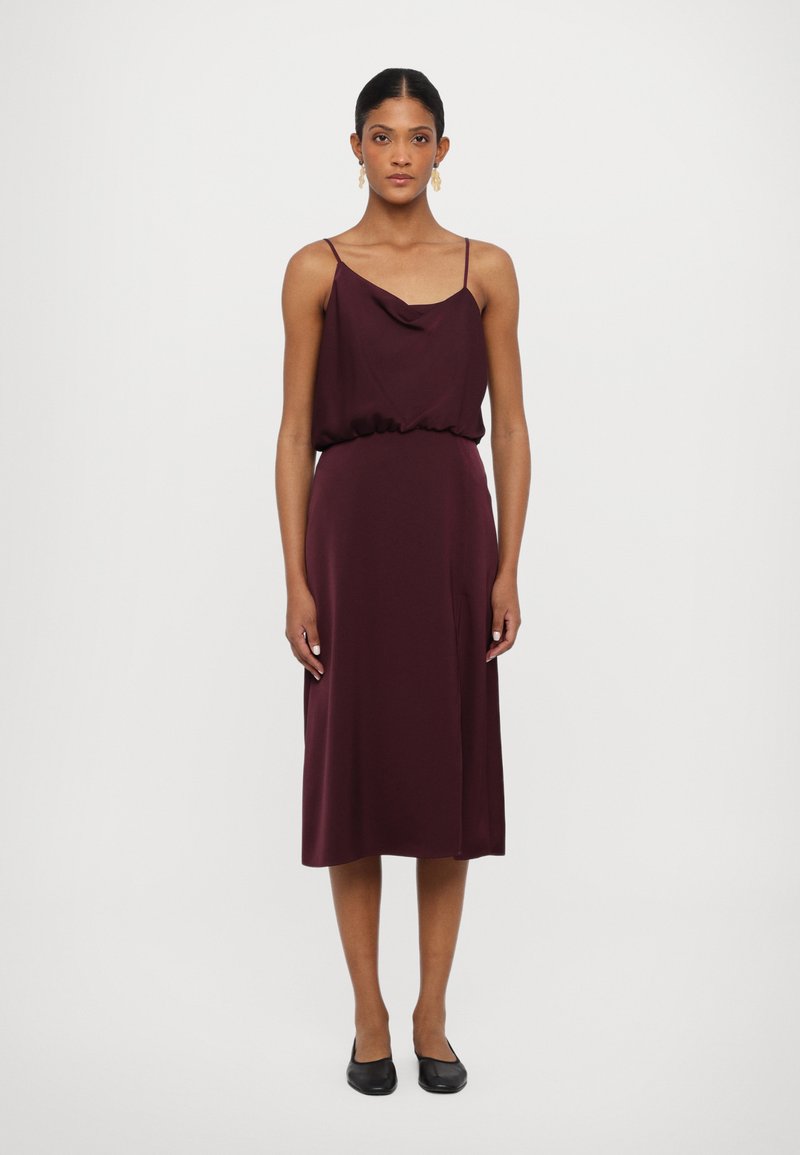 Rochie midi bordeaux cu guler înfășurat, talie elastică și bretele subțiri. Material neted și ușor. Modelul poartă balerini negri.