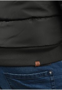 Schwarze Steppjacke mit geripptem Stoff am Saum und einem braunen Leder-Logoetikett. Getragen über blauen Jeans mit sichtbaren Taschen-Details.