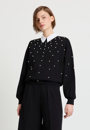 Femme portant un pull noir avec des perles blanches dispersées sur une chemise blanche à col et un pantalon noir taille haute sur un fond neutre.