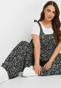 Svart blommig jumpsuit med vita blommor, justerbara band och en avslappnad passform, kombinerad med vita sneakers på en sittande figur.