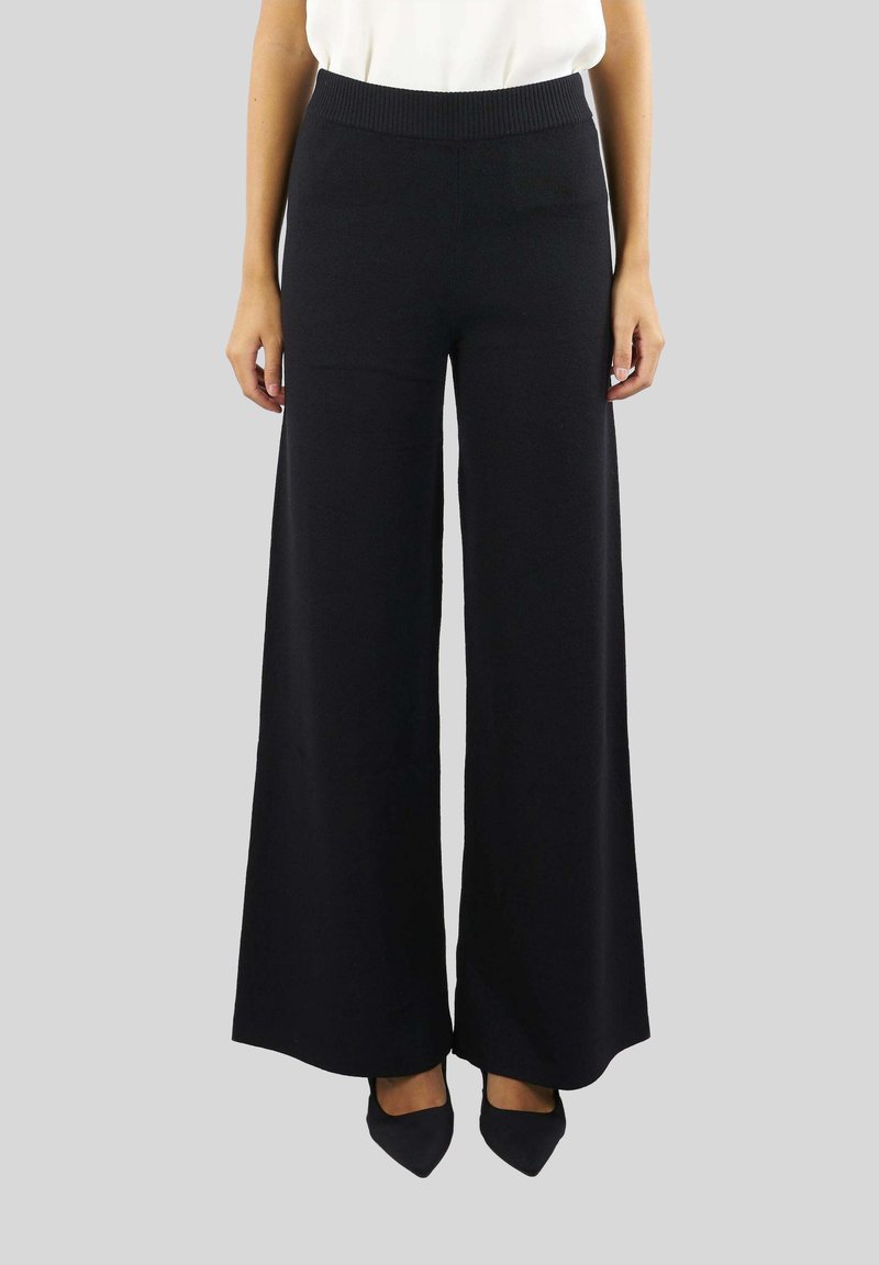 Pantaloni neri a gamba larga realizzati in un materiale morbido, con una vita a costine e una texture liscia, abbinati a scarpe nere a punta.