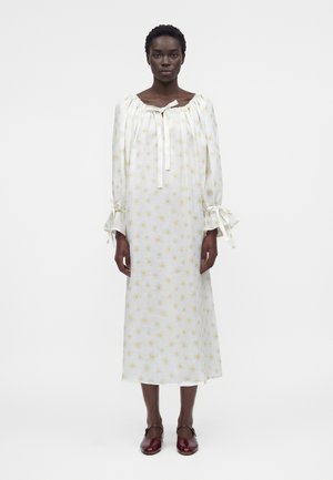 Kvinde iført en hvid lang kjole med gule blomsterprint, indsamlet halsudskæring med bindebånd, sløjfedetaljer på ærmerne og mørkerøde Mary Jane-sko.