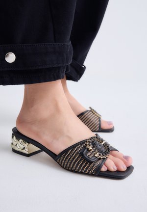 Primer plano de pies con sandalias negras tejidas sin cierre con hebilla decorativa en tono dorado y tacones bajos metálicos esculpidos en bloque.