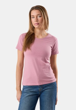 Femme aux longs cheveux châtain clair portant un t-shirt rose pâle uni à manches courtes et un jean bleu, debout devant un fond blanc uni.