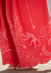 Vêtement en tissu rouge orné de broderies blanches représentant des feuilles, un singe, des étoiles et des motifs circulaires, avec un ourlet festonné et de petits points décoratifs.