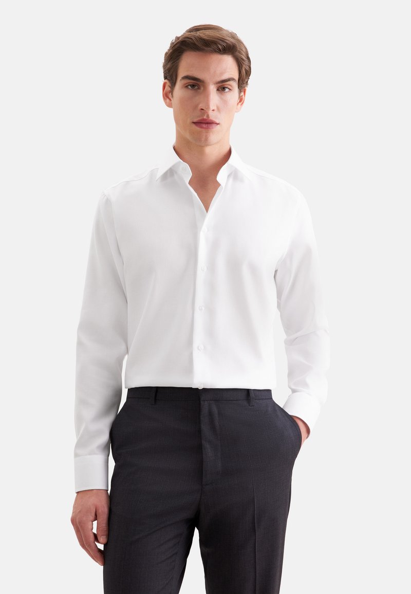 Homme portant une chemise blanche à manches longues boutonnée et un pantalon sombre, debout avec une main dans la poche sur un fond uni.