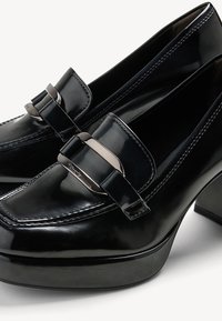 Mocassins en cuir verni noir au design épuré, avec un accent de lanière métallique et un talon épais. Texture lisse et détails piqués.