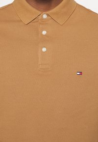 Polo en coton marron avec une finition texturée. Doté d'une patte de boutonnage et d'un petit logo brodé en rouge, blanc et bleu sur la poitrine.