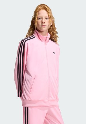 Young person with long curly red hair wearing a pink Adidas tracksuit with black stripes on sleeves and pants against a plain background.