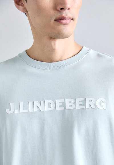 Hele sinise puuvillase T-särk, millel on ümarad kraed ja ees suur valge trükitud logo "J.LINDEBERG". Sile tekstuur.