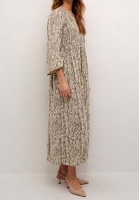 Femme portant une robe longue  à motif tourbillonnant beige et marron avec des manches trois-quarts et des chaussures à talons bas beige à l'arrière.