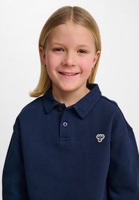 Mörkblå polo med ett texturerat mönster, med en tvåknäppningsplacket och en ljusfärgad emblem på vänster sida.