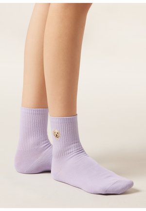 Beine mit helllila gerippten Socken, die ein kleines besticktes Golden-Retriever-Hundegesicht am äußeren Knöchel zeigen.