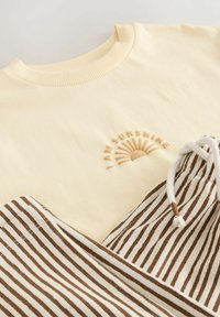 Cremefarbener Sweatshirt mit geripptem Kragen und dem Schriftzug "I AM SUNSHINE" sowie Sonnenmotiv, kombiniert mit braunen und weiß gestreiften Hose mit Kordelzug.