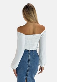 Haut blanc à épaules découvertes avec texture smockée et manches longues, associé à une jupe en jean taille haute avec une fermeture éclair sur l'arrière et une fente.